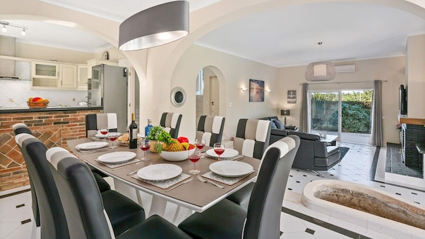 A modern dining area with a brick accent wall, a large table set for six, and a view of the kitchen and outdoor patio. - Villa Casa do Mel by Villa Plus (Loule)