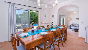 Een eetgedeelte met een houten tafel gedekt voor vier personen, een kroonluchter en uitzicht op een zwembad en groen buiten.