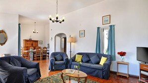 Ein Wohnzimmer mit zwei Sofas, einem Couchtisch und einer TV-Konsole. Eine Essecke mit Tisch und Stühlen ist vorhanden, und durch einen Bogen ist die Küche zu sehen.