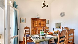 Ein Essbereich mit einem Holztisch, gedeckt für vier Personen, umgeben von Korbstühlen mit blauen Kissen. Im Hintergrund steht eine Holzkommode mit dekorativer Oberseite.