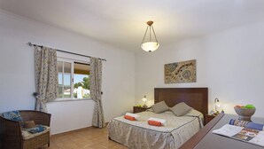 Een slaapkamer met een bed, een bureau, een stoel, een raam met gordijnen, een hanglamp en een schilderij aan de muur.