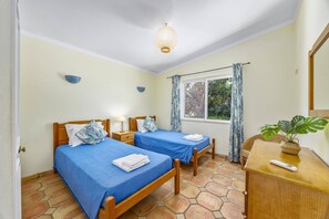 A room with two beds, a window with curtains, a bedside table with a lamp, and a plant on a wooden dresser. - Villa Flor by Villa Plus (Guia)