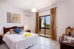 Un dormitorio con cama, mesita de noche, lámpara, ventana con cortinas, un cuadro en la pared y vistas al exterior.