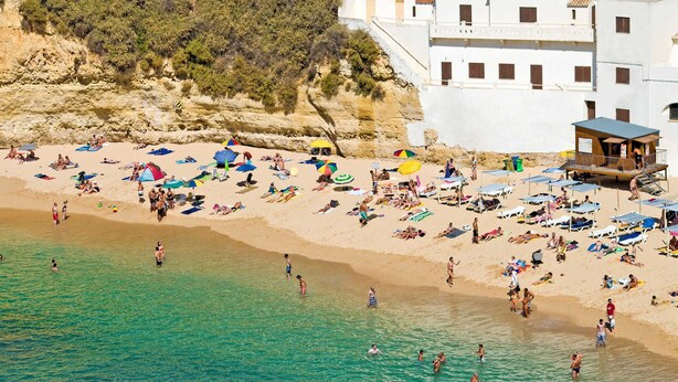 A beach with sunbathers and umbrellas, a lifeguard tower, and a building with balconies. - Villa Lisson Grove by Villa Plus (Carvoeiro)