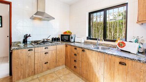 A kitchen with wooden cabinets, a stainless steel range hood, a microwave, a toaster, and a window with a view of greenery. - Villa Lisson Grove by Villa Plus (Carvoeiro)