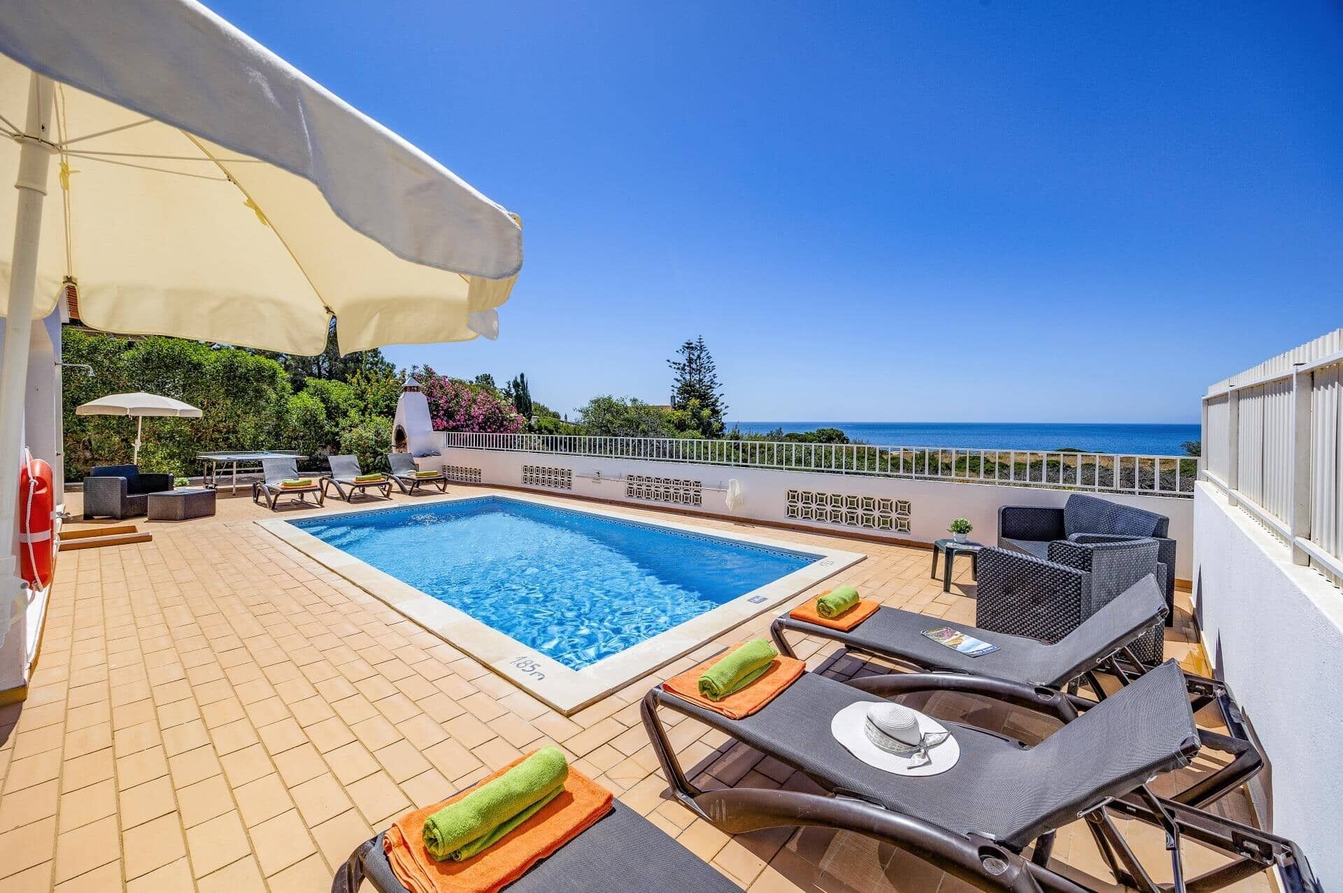 Área de piscina no terraço com vista para o mar, cadeiras de descanso e guarda-sóis.