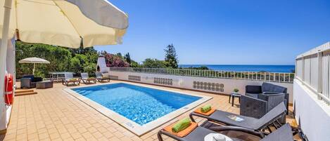 A rooftop pool area with a view of the sea, lounge chairs, and umbrellas.