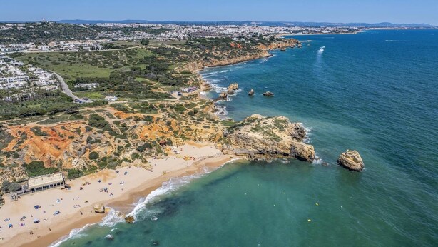 Eine Luftaufnahme einer Küstenlandschaft mit Strand, Felsen und einer Stadt im Hintergrund.