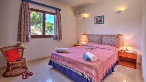 Quarto com uma cama, duas mesas de cabeceira, uma cadeira de vime, uma janela com cortinas e um aparelho de ar condicionado.