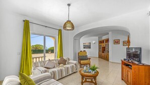 Uma sala de estar ampla com sofá, poltrona e uma pequena mesa redonda com uma planta. Uma televisão está sobre um móvel de madeira, e há um quadro na parede.