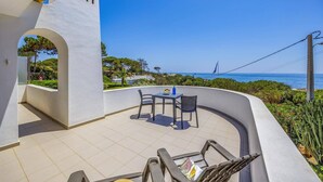 Uma varanda com mesa e cadeiras, vista para o mar, e um edifício branco com janelas em arco.
