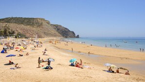 Ein Strand mit badenden und sonnenden Menschen, ein felsiger Klippenhügel im Hintergrund und ein klarer, blauer Himmel.