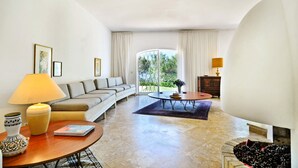 Ein geräumiges Wohnzimmer mit Sofa, Couchtisch und Esstisch. Es gibt ein großes Fenster mit Vorhängen, eine Lampe und eine Vase mit Blumen.