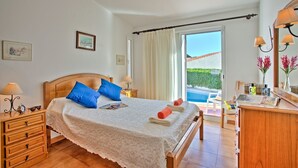 Un dormitorio con una cama de madera, dos almohadas azules, una colcha blanca, una mesita de noche con lámpara, un aparador, una ventana con cortinas y vistas a una piscina.