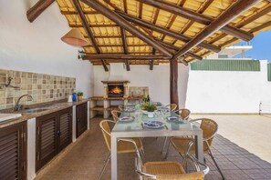 Une terrasse extérieure avec une table à manger pour quatre personnes, entourée de chaises en osier. On aperçoit un barbecue intégré à l’arrière-plan, et un pare-feu en carreaux fait partie de l’espace cuisine.