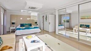 Ett modernt hotellrum med en stor säng, en sittgrupp med soffa och soffbord, en glasdörr som leder ut till en balkong och en stor spegel som reflekterar rummet.