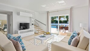 Ett modernt vardagsrum med tv, soffa, soffbord och utsikt över en pool och ett utomhusområde för måltider.
