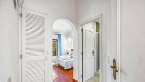 Ein Flur, der zu einem Schlafzimmer mit Bett, Nachttisch und einem Fenster mit Blick auf Grünfläche führt.