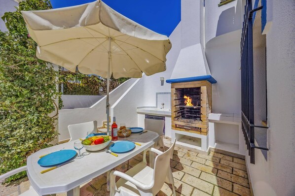Une terrasse avec une table dressée pour deux personnes, un barbecue et un mur blanc orné de motifs bleus.