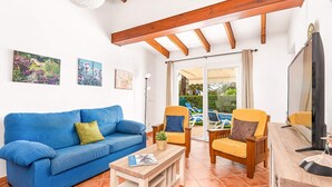 Een woonkamer met een blauwe bank, houten stoelen, een houten salontafel en een televisie. Er hangen schilderijen aan de muur en er is uitzicht op het exterieur door een glazen deur.