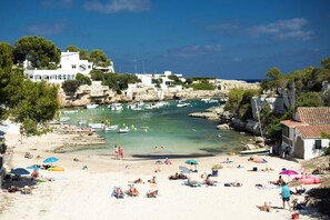 A beach with numerous sun loungers and umbrellas, boats in the water, and buildings in the background. - Villa Beltran by Villa Plus (S'Algar)