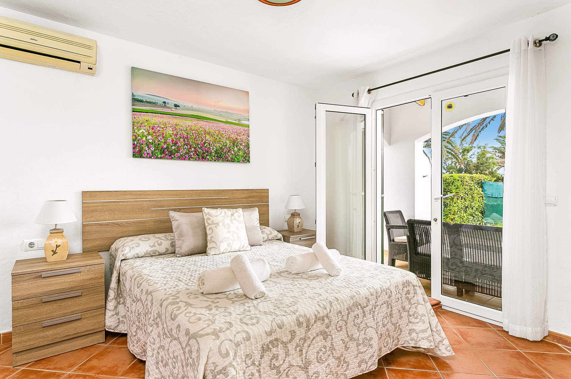 A bedroom with a bed, a nightstand, a lamp, a painting, and a sliding glass door to a balcony.