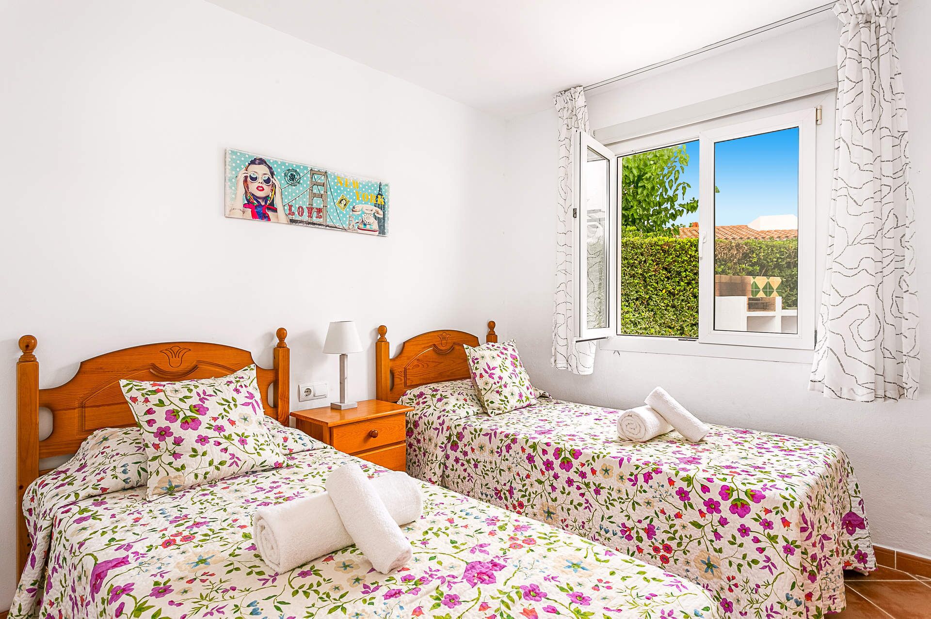 A bedroom with two wooden beds, floral bedding, a bedside table with a lamp, a painting on the wall, and a window with a view of greenery.