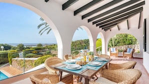 Un coin repas avec des meubles en osier, une table dressée pour deux personnes et une vue sur la mer et la verdure.