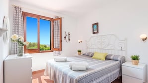 Une chambre à coucher comprenant un lit, deux tables de chevet, une commode, une fenêtre avec des rideaux, un tableau accroché au mur et un luminaire fixé au mur.