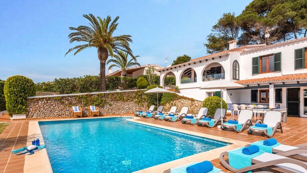 Une piscine entourée d’un mur de pierre et de palmiers, avec des chaises longues et des parasols.