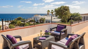 A balcony with wicker furniture, a table with a bottle and glass, and a view of the sea and buildings.