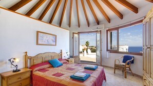 A bedroom with a wooden ceiling, a bed with a red and orange quilt, a nightstand with a lamp, a chair with a blue cushion, and a view of the sea through a window.