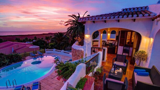 A pool area with lounge chairs, a dining area with tables and chairs, and a view of the ocean at sunset. - Villa Corina by Villa Plus (Alayor)