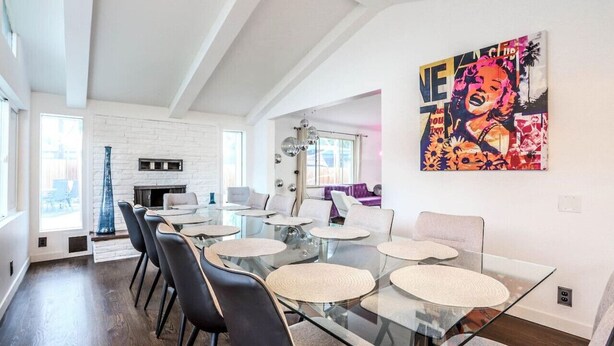 Un comedor moderno con una mesa de vidrio y sillas negras, una chimenea de ladrillo blanco y un cuadro de arte pop vibrante en la pared.