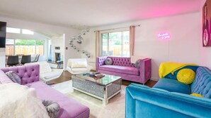 Una sala de estar moderna con un sofá morado, un sofá azul y un sillón blanco. Hay una mesa de centro de vidrio y una chimenea al fondo.