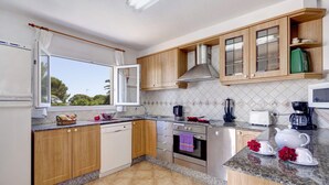 A modern kitchen with wooden cabinets, a dishwasher, an oven, and a window with a view of trees. - Villa Fernando by Villa Plus (S'Algar)