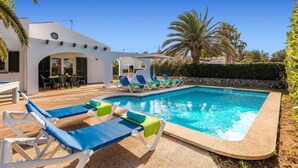 A swimming pool area with lounge chairs and a patio table, surrounded by palm trees and a white building. - Villa Jessica by Villa Plus (Cap d'Artrutx)