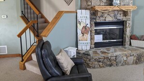A living room with a stone fireplace, a wooden staircase, a black leather sofa, and a decorative sign. - Beautiful Townhouse in The Villages of 7 Springs
 (Champion)