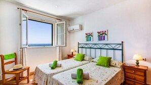A room with two beds, a window with an open curtain, a chair, a wooden dresser, and a wall with two floral paintings. - Villa Laurel by Villa Plus (Alayor)