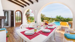 Un área de comedor al aire libre con una mesa para cuatro, con vistas a una piscina y un jardín.