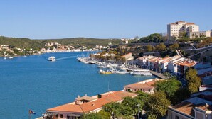 A coastal town with a marina, buildings with red-tiled roofs, and a prominent hill with a building on top. - Villa Mirador by Villa Plus (Cala en Porter)