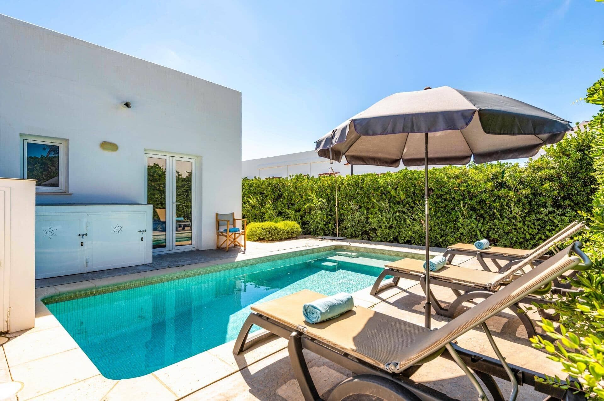 A modern outdoor pool area with lounge chairs and an umbrella, surrounded by a neatly trimmed hedge and a white building.