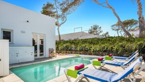 Un área de piscina con sillones de descanso y un edificio blanco con una puerta y ventanas.