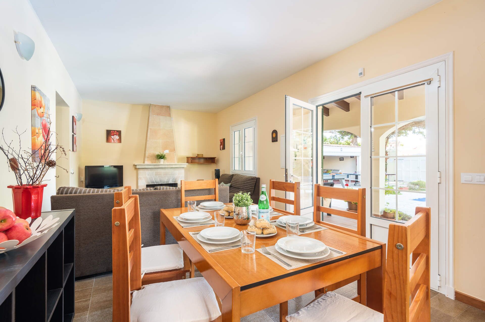 Un comedor con una mesa de madera dispuesta para seis personas, rodeada de sillas. Hay un sofá y un televisor al fondo.