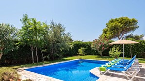 A rectangular swimming pool with a stone border, surrounded by a garden with various trees and shrubs. There are lounge chairs with blue cushions and a sun umbrella.
