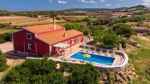 En rød villa med swimmingpool, omgivet af grøn vegetation og bakker.