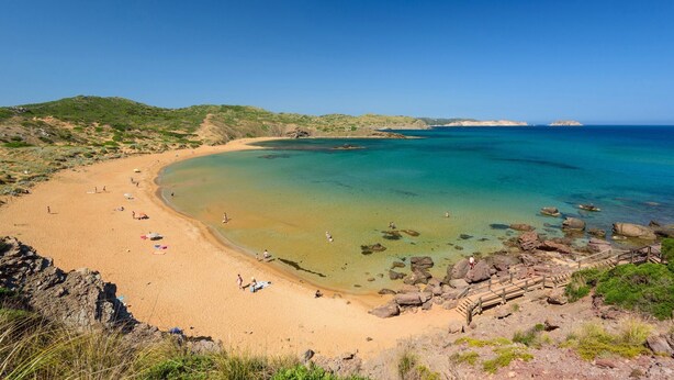 En strand med klart turkis vand, sandet bredde og klippeformationer.