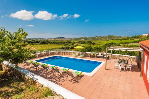 Una piscina rectangular con pared de piedra y solárium con baldosas, rodeada de sillas de descanso y una pérgola con mesa y sillas.