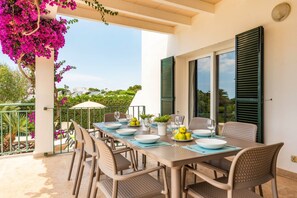 An outdoor dining area with a table set for four, surrounded by chairs, with a view of greenery and buildings in the background.