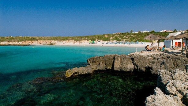 Cena de praia costeira com águas turquesa cristalinas, formações rochosas e alguns guarda-sóis de praia.